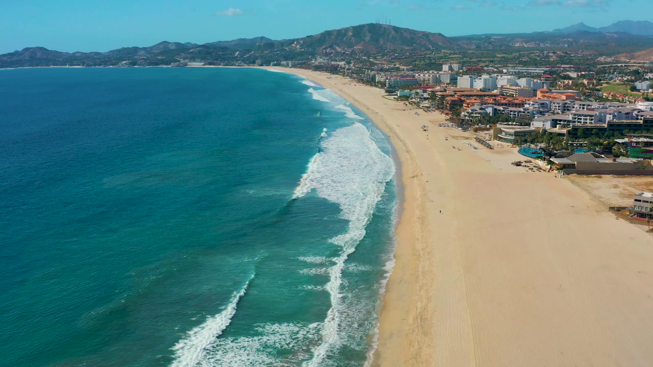 Aerial beach view with rolling surf, open sand, coastal buildings, and mountains.