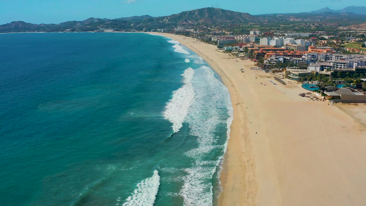 White wave foam advances along the shoreline beside a broad sand beach and coastal buildings.