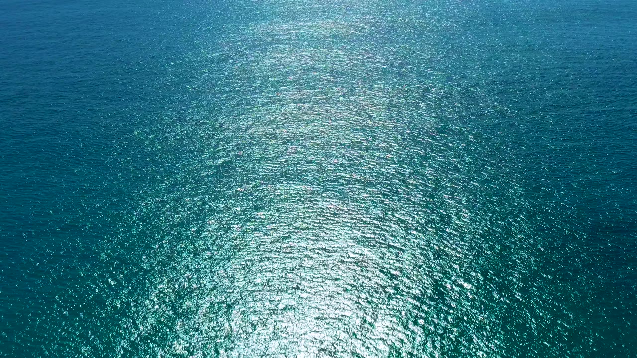 Aerial view of blue and teal ocean water in San Jose Del Cabo with a broad sunlight reflection near the lower center and no visible people.