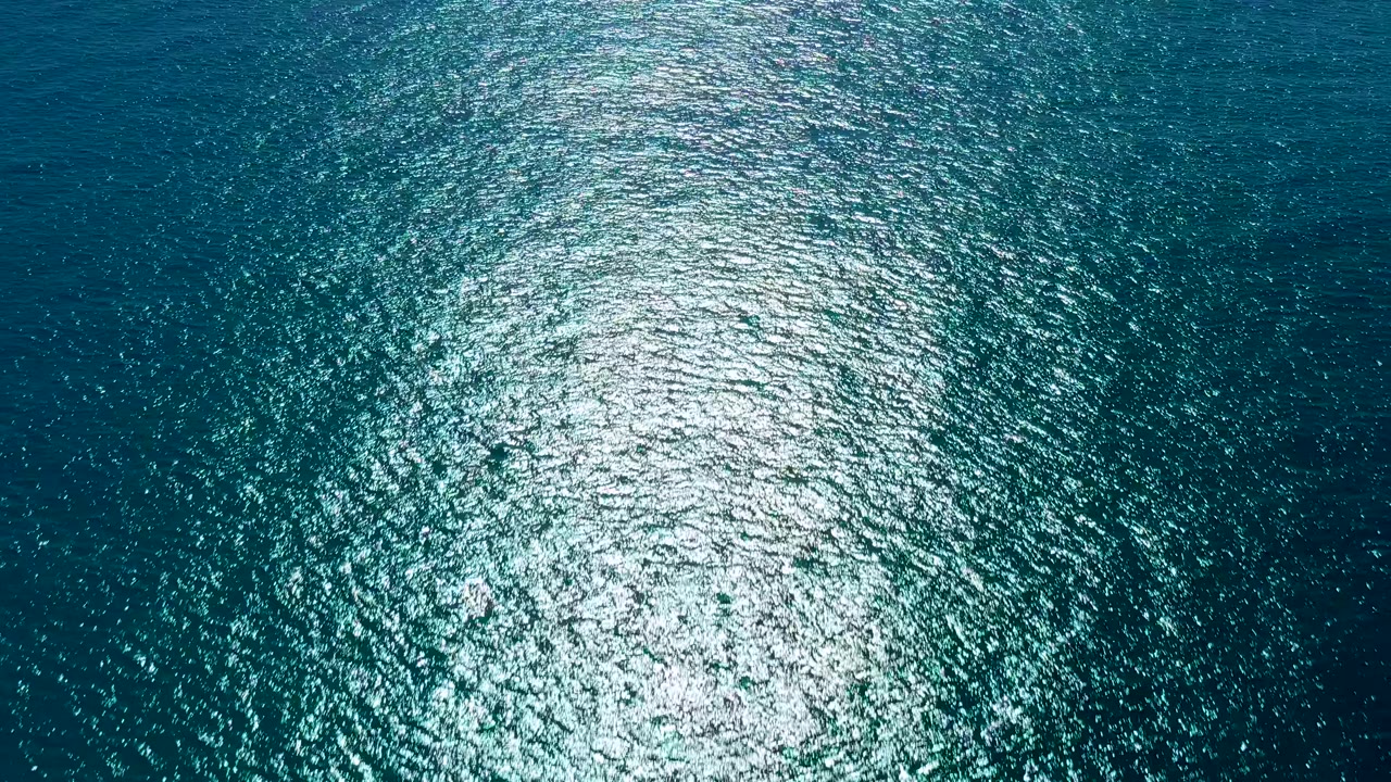 Overhead view of rippling teal ocean in San Jose del Cabo with a bright sunlight reflection near the lower left, no visible people.