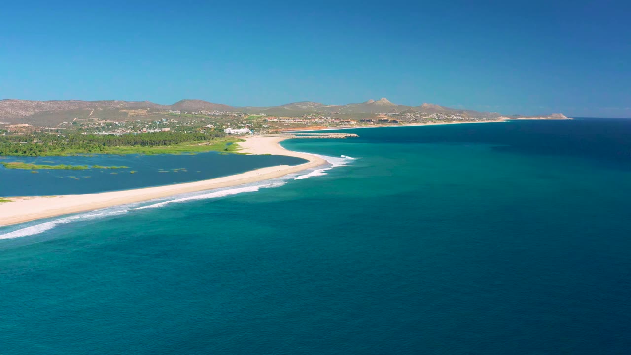 San Jose del Cabo aerial view with white surf lines advancing along the pale sand, deep blue water offshore, green shoreline vegetation, and no visible people.