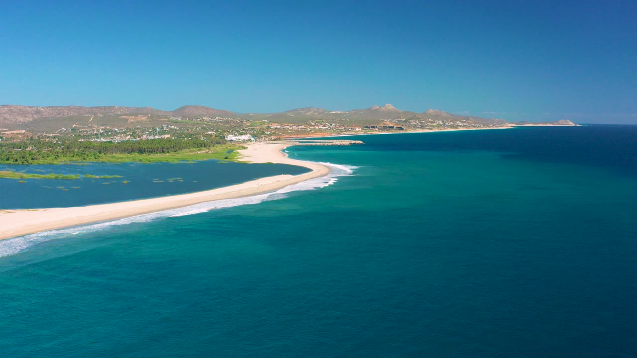 Aerial frame over San Jose del Cabo showing a pale sandy spit between turquoise ocean and a greenish inlet, with low hills under clear blue sky and no visible people.