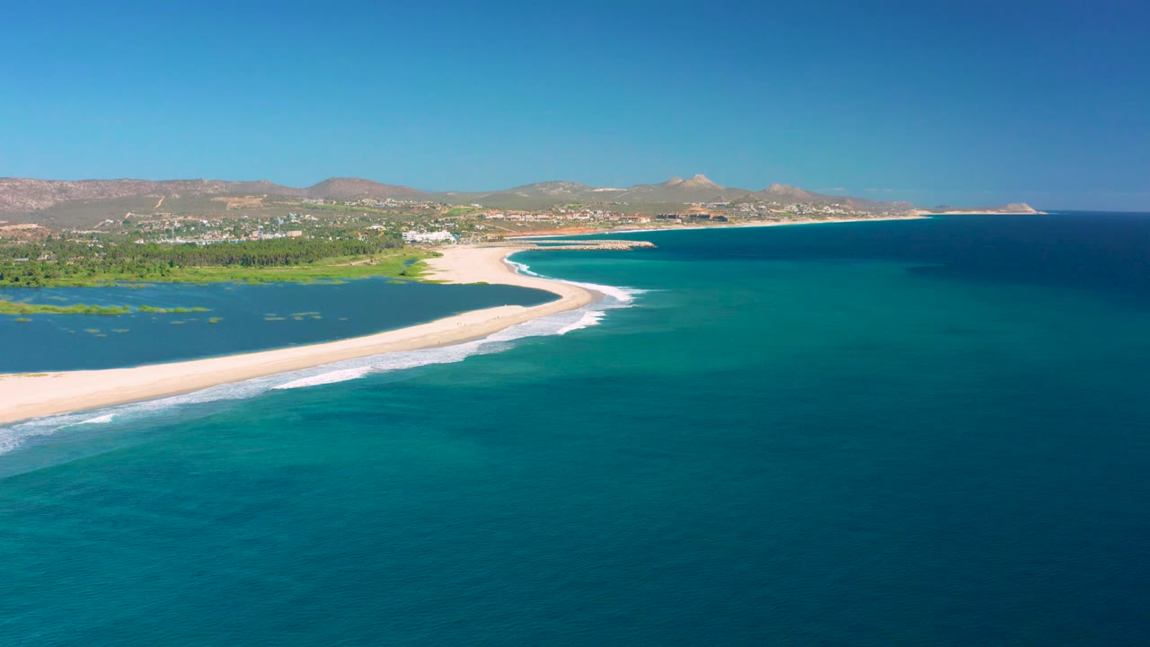 Aerial coastline with sandy spit, green inlet, turquoise ocean, and low mountains.