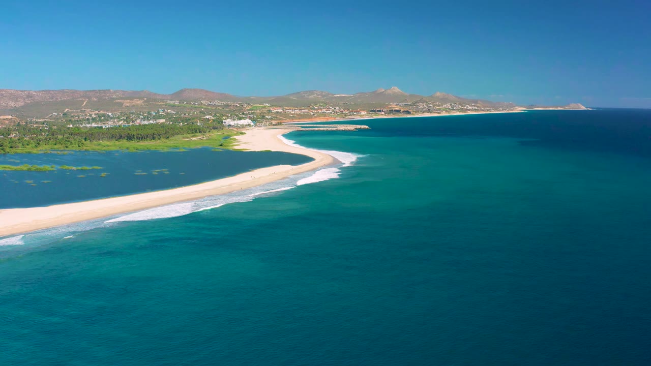 San Jose Del Cabo coastline seen from above, with the sand spit crossing the frame, darker offshore water to the right, and no visible people.