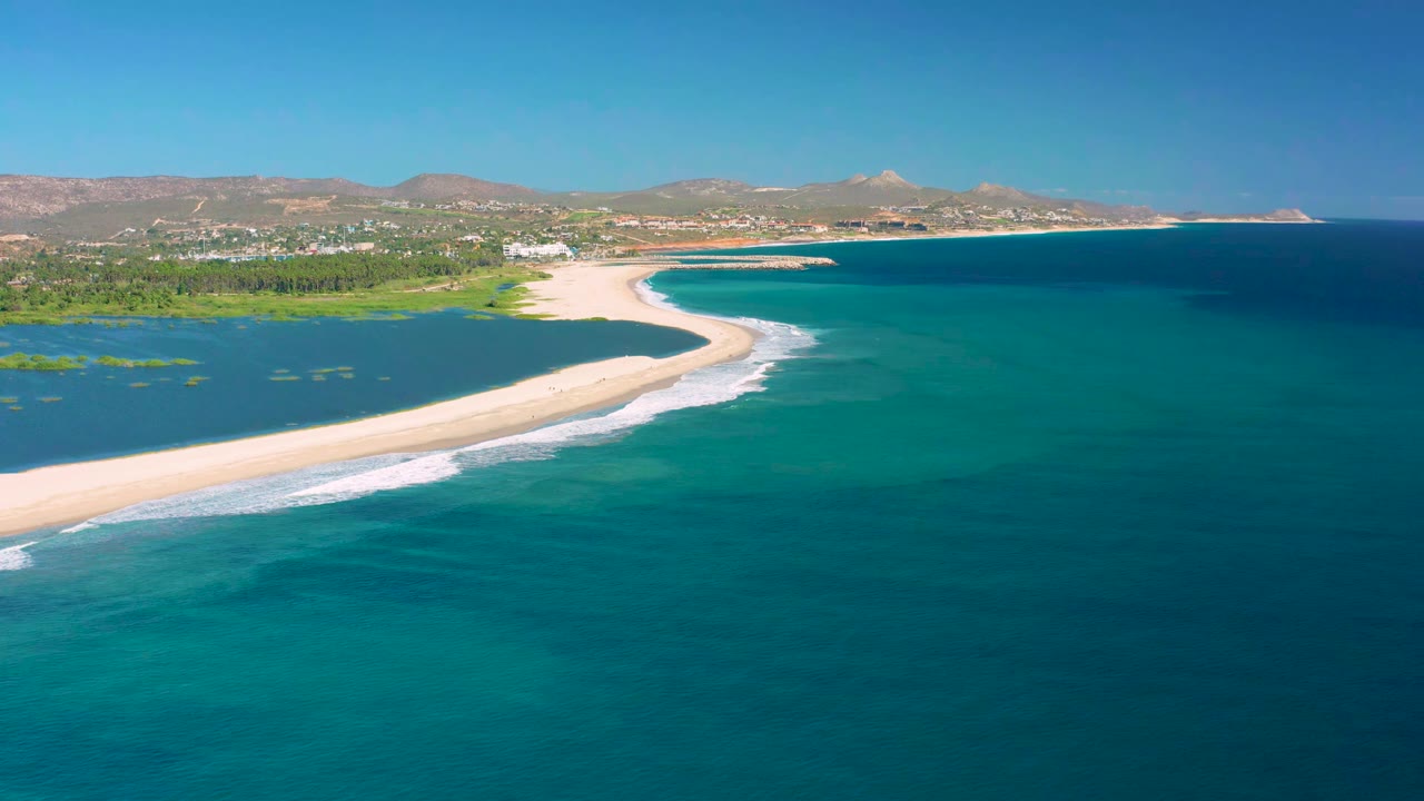 Aerial view in San Jose Del Cabo with the curved sandbar more centered, white waves breaking along the ocean side, and no visible people.