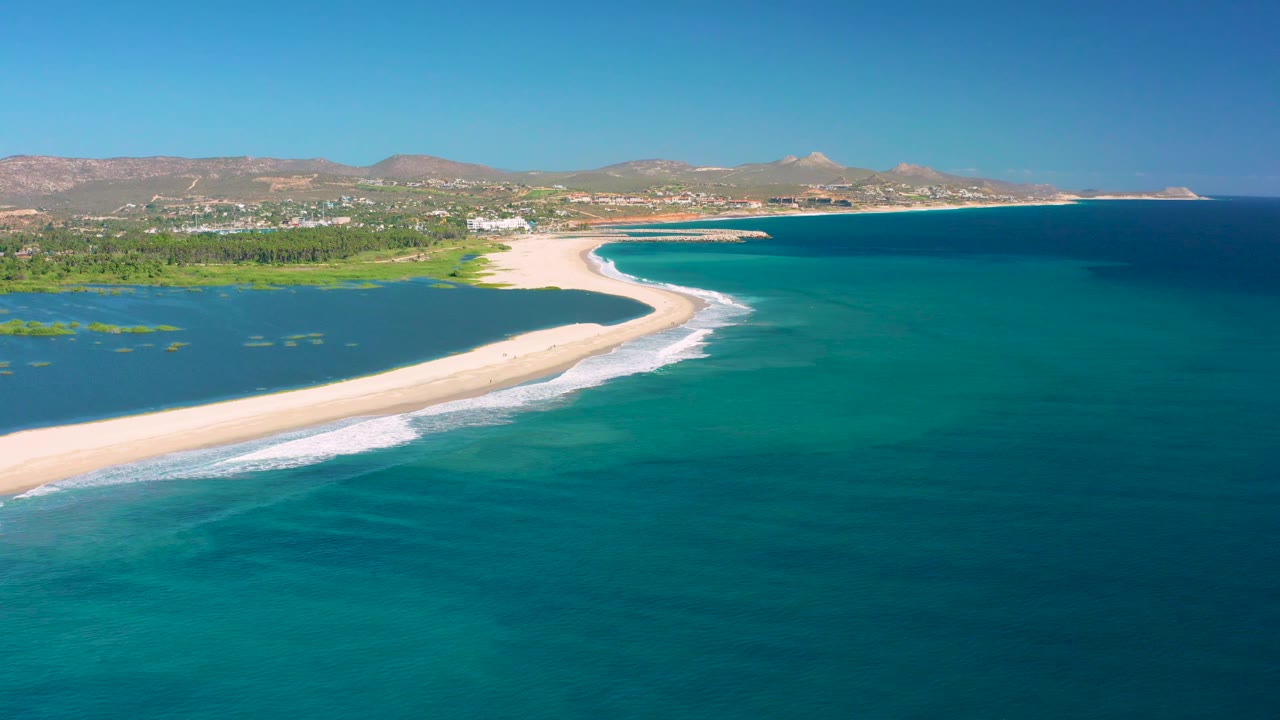 Mid-clip aerial view over San Jose Del Cabo showing lagoon patches at left, a long sandy barrier, rolling white surf, and no visible people.