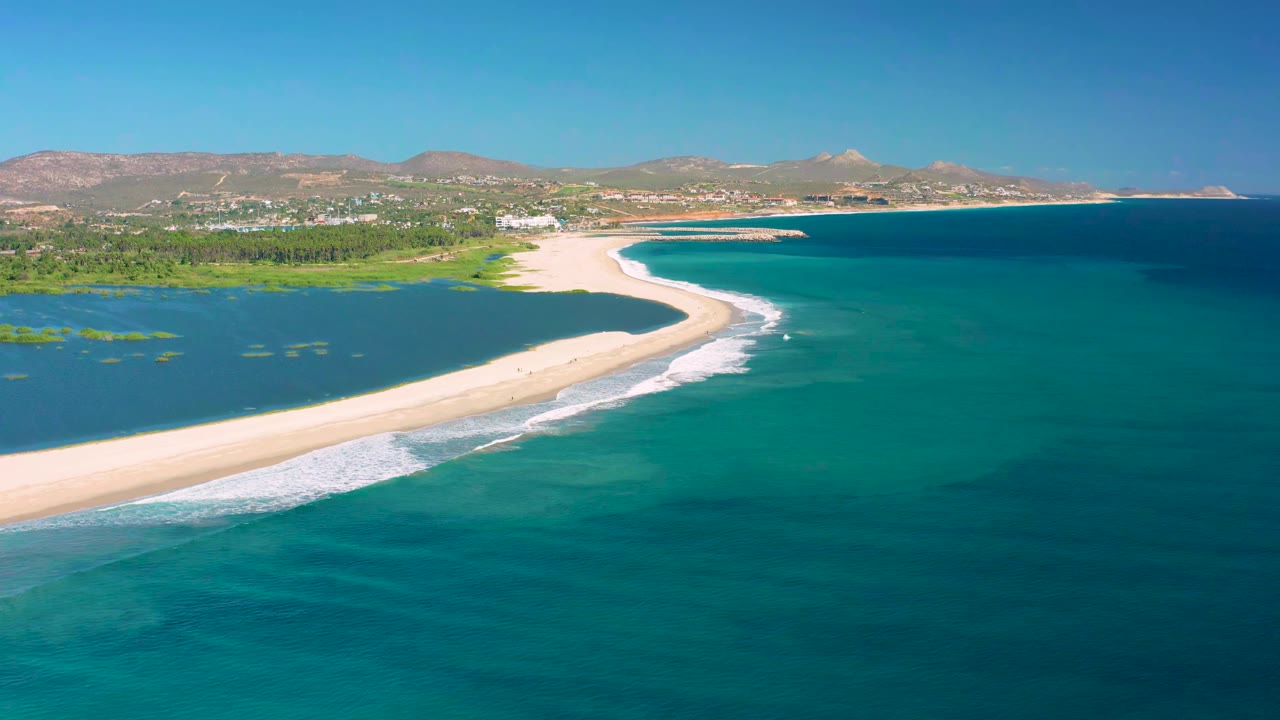 Aerial view in San Jose Del Cabo with the curving sandbar slightly more centered, white foam along the ocean side, green lagoon water to the left, and no visible people.