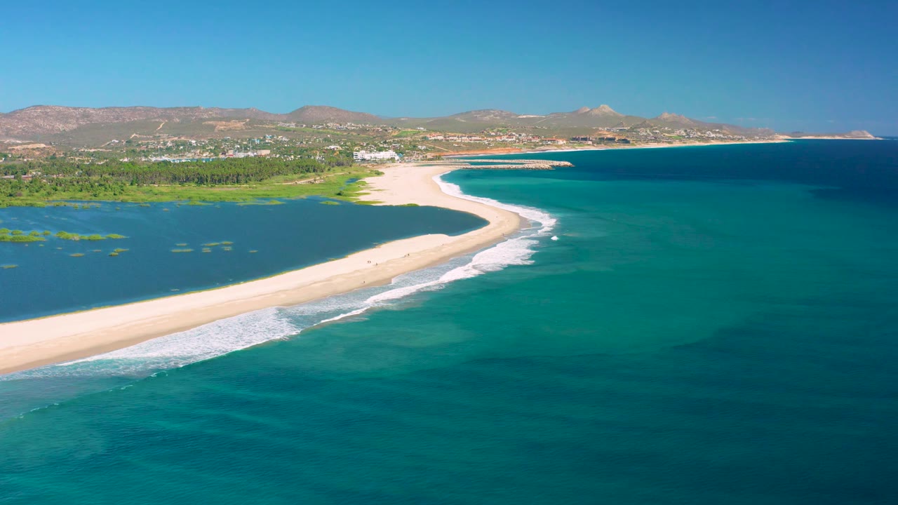 Wide San Jose Del Cabo aerial frame with the lagoon and green shoreline on the left, a light sand spit across the middle, deeper ocean on the right, and no visible people.