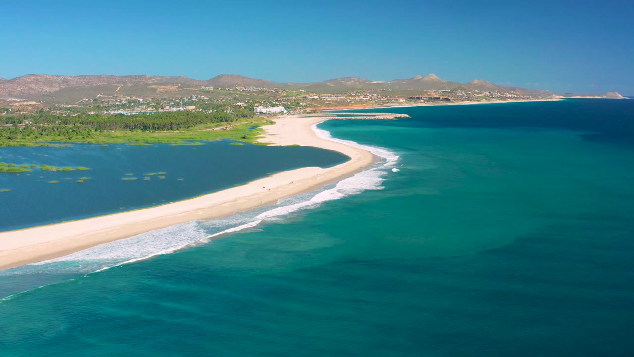 San Jose Del Cabo aerial still with the sand spit running diagonally across the foreground, calm lagoon water to the left, ocean surf on the right edge, and no visible people.