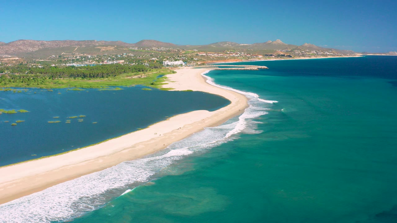 Aerial view in San Jose del Cabo showing a pale sandy barrier island between a green-blue lagoon and turquoise ocean, with white surf at the lower edge and no visible people.