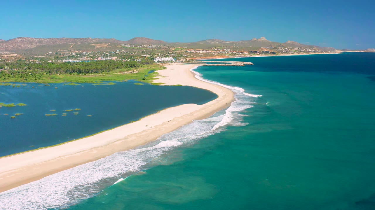 Aerial frame over San Jose del Cabo with the sandbar curving from lower left toward the town edge, lagoon water on the left, ocean water on the right, and no visible people.