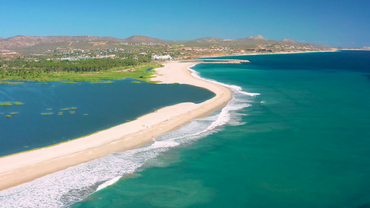 Wide aerial view in San Jose del Cabo where the sandy spit separates dark lagoon water from clear turquoise ocean, with foam bands along the beach and no visible people.