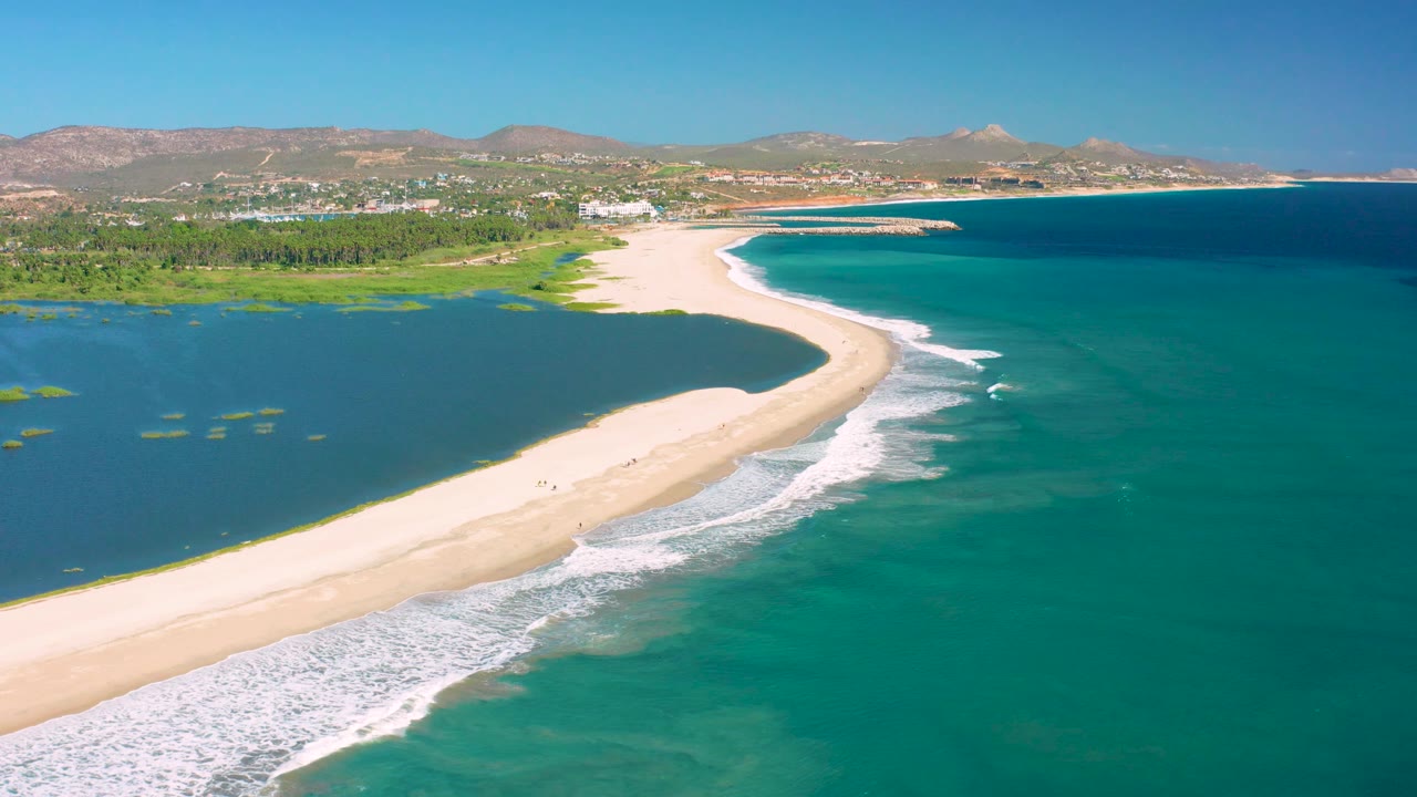 Aerial coastline with pale sand, lagoon water, surf, and distant buildings.