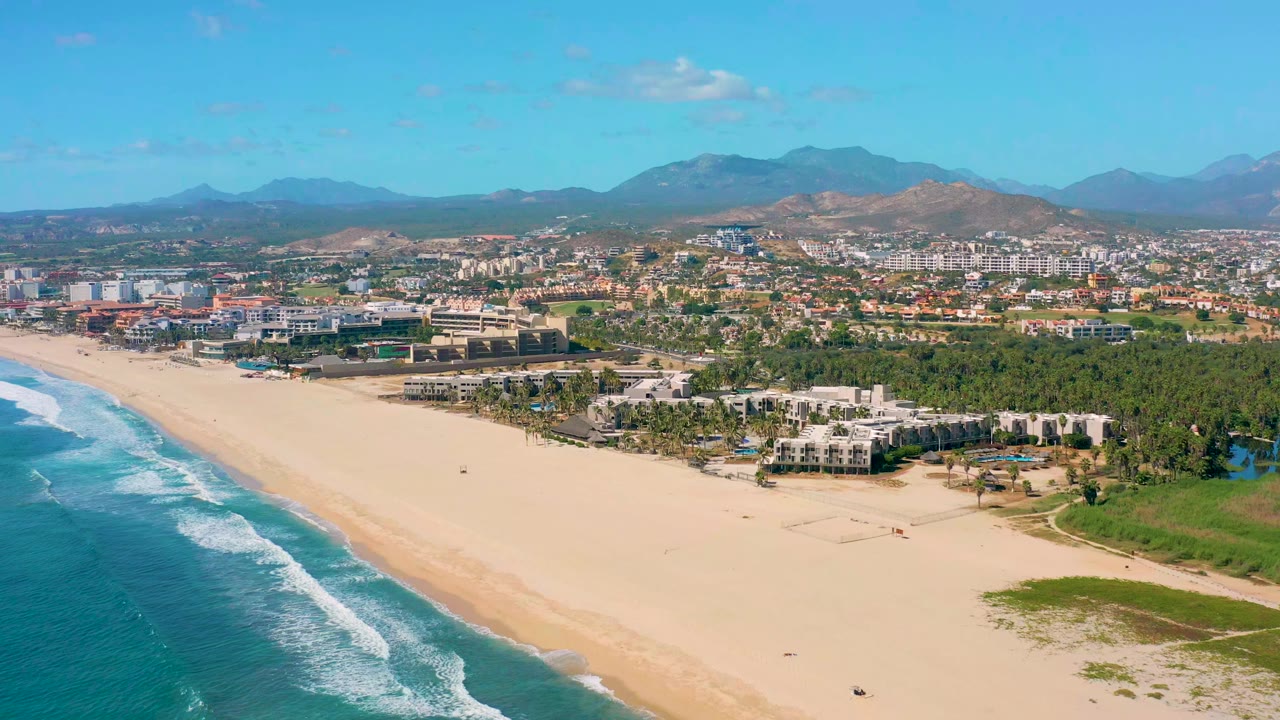 Elevated San Jose del Cabo beach frame with white wave lines closer to shore, pale sand across the center, coastal buildings behind the beach, mountain ridges in the distance, and no visible people.