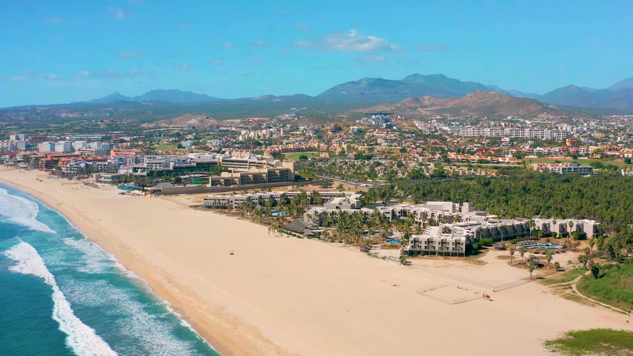 Second aerial frame in San Jose Del Cabo, Mexico, with white surf moving up the left side, open tan sand in front, clustered buildings and palms behind it, mountains on the horizon, and no visible people.