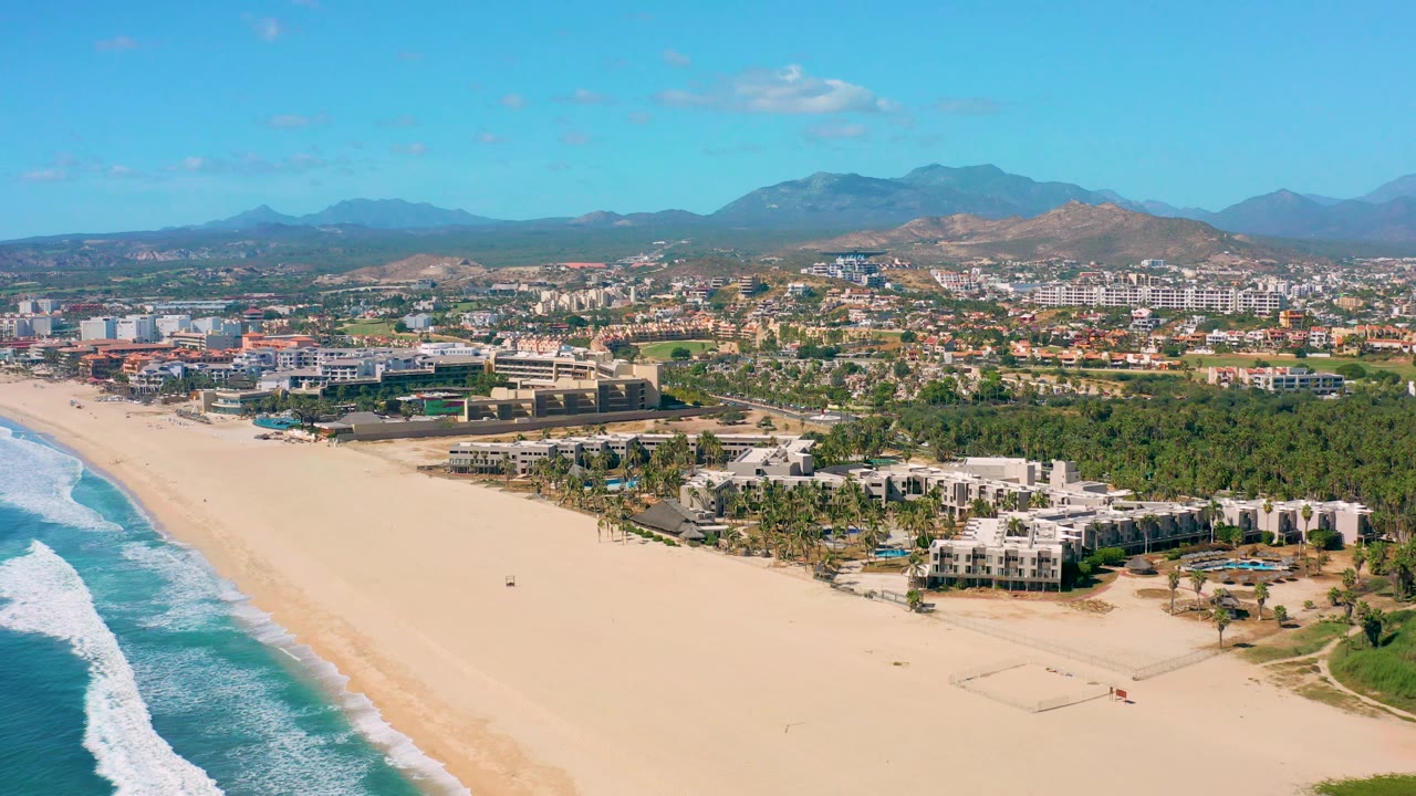 Fourth frame in San Jose Del Cabo, Mexico, places the surf strip along the left border, the sandy beach across the lower frame, white buildings and palms in the midground, town blocks beyond, mountains in back, and no visible people.