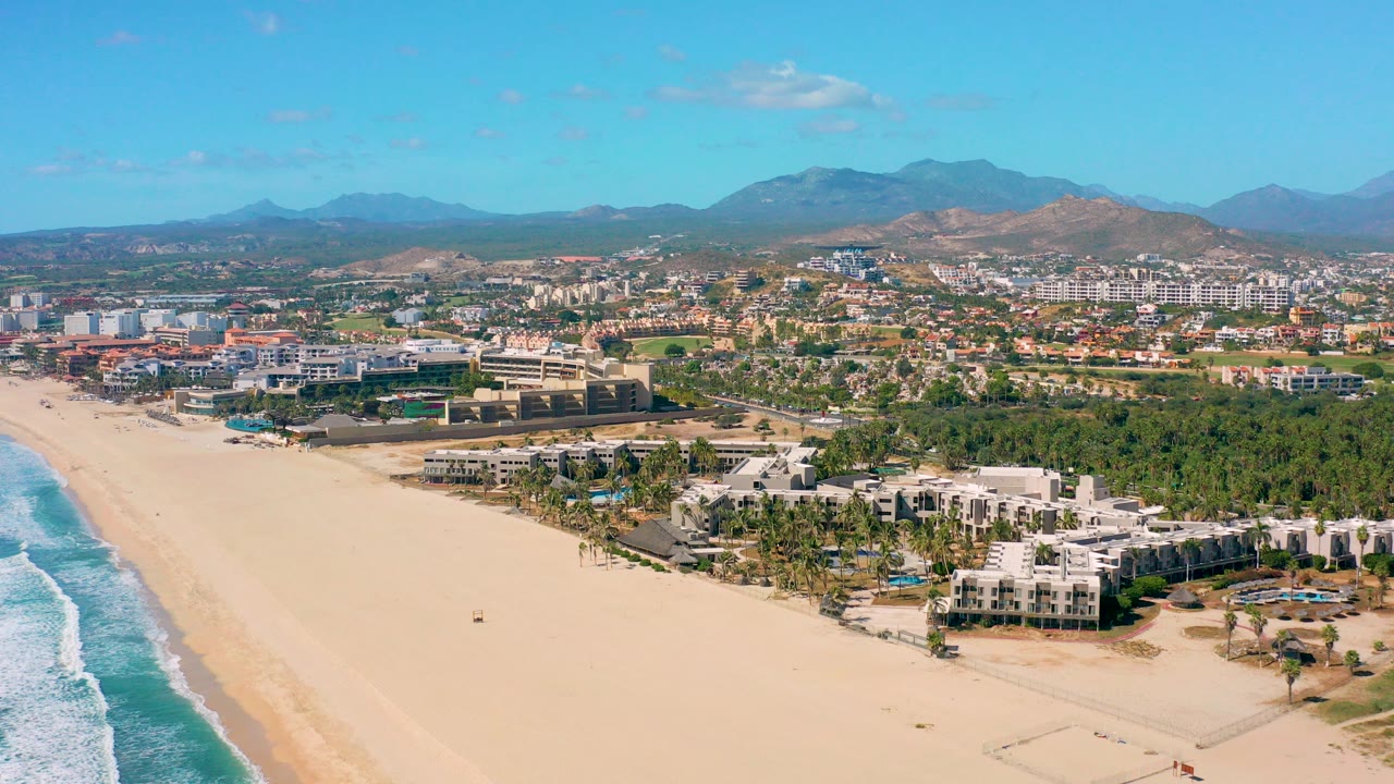 High aerial view over San Jose del Cabo beach with blue surf at left, pale sand in front, palm-lined coastal buildings, inland city blocks, distant mountains, and no visible people.