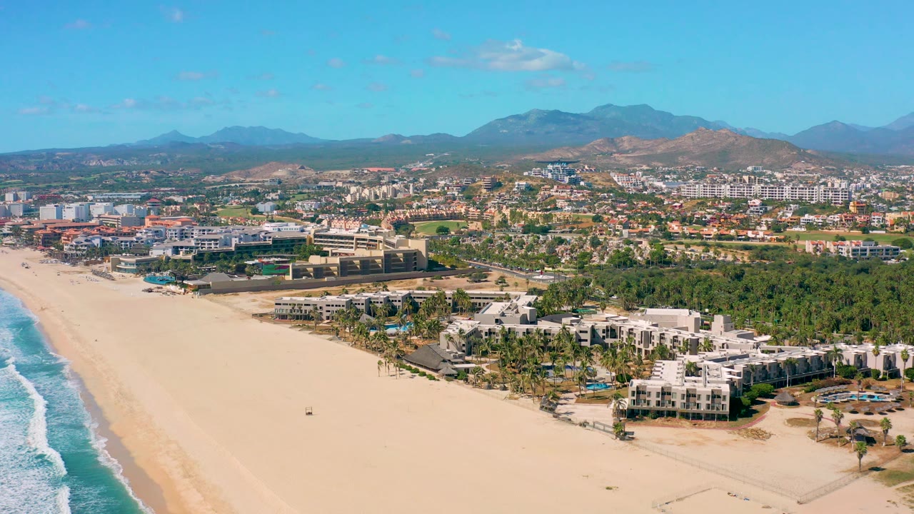 San Jose del Cabo coastline from above, with the ocean edge along the left, an empty-looking sandy beach across the foreground, white coastal buildings at lower right, green palms, city blocks, hazy mountains, and no visible people.