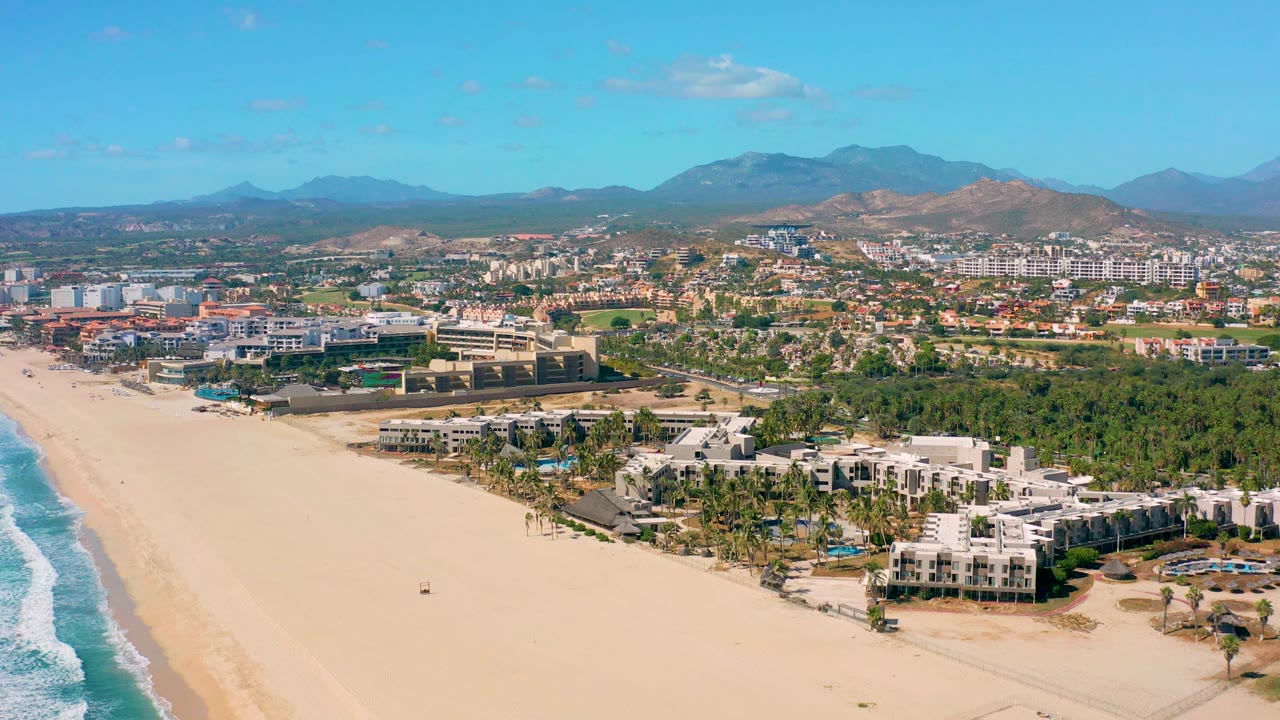 Elevated aerial view in San Jose del Cabo with pale sand and surf at left, coastal hotel-style buildings and palm trees at lower right, inland city development, mountain ridges, scattered clouds, and no visible people.