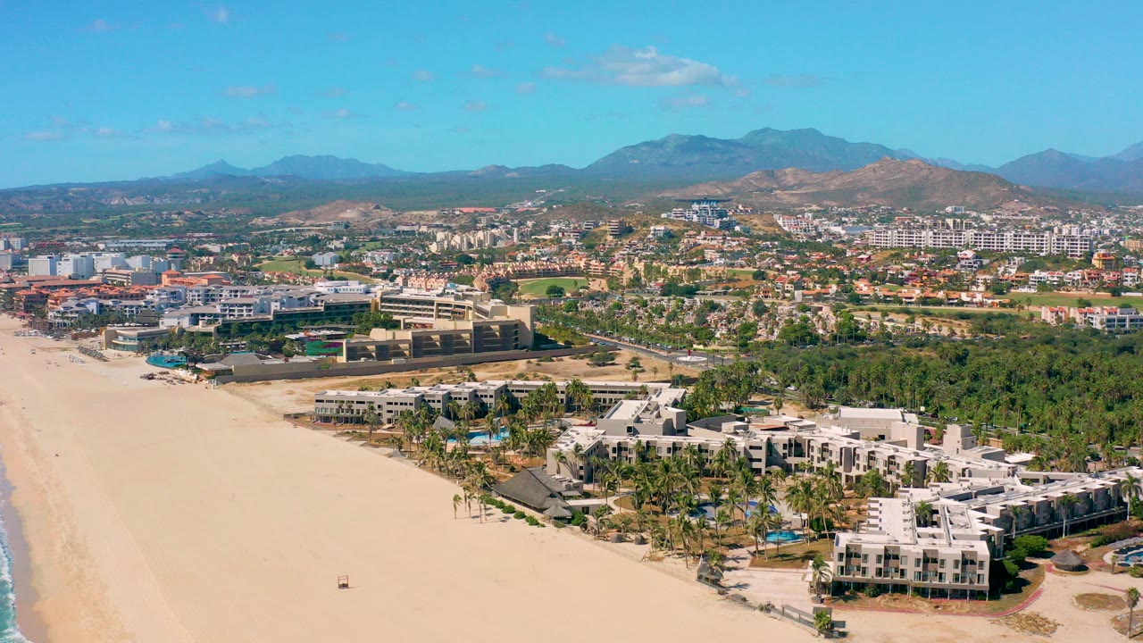Opening aerial frame over San Jose Del Cabo with a wide sandy beach at left, white resort buildings near the lower edge, town development beyond, mountains on the horizon, and no visible people.