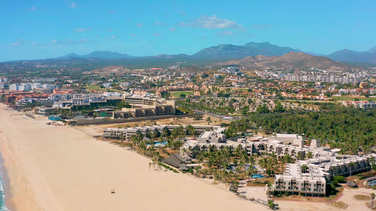 Aerial view in San Jose Del Cabo showing the beach running up the left side, a white resort complex and palms along the lower right, distant buildings, mountains, and no visible people.