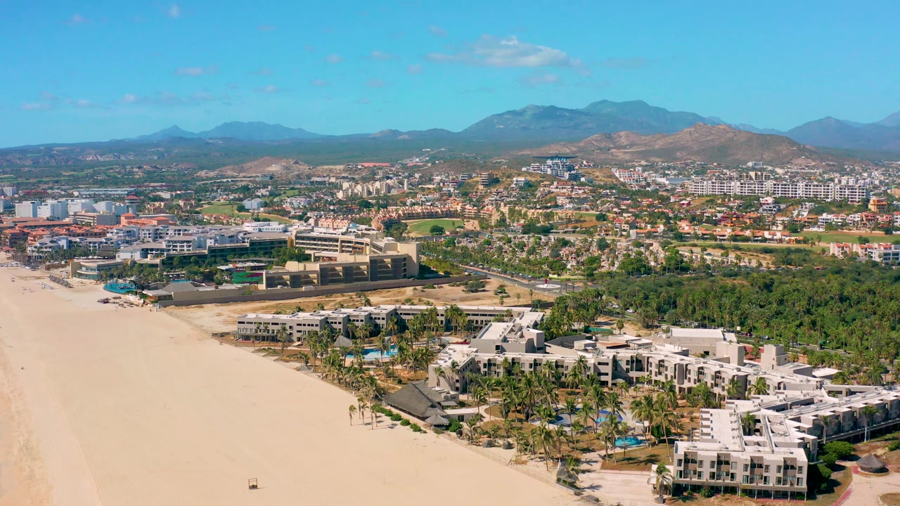 Aerial beach and resort view with palms, low buildings, and distant hills.