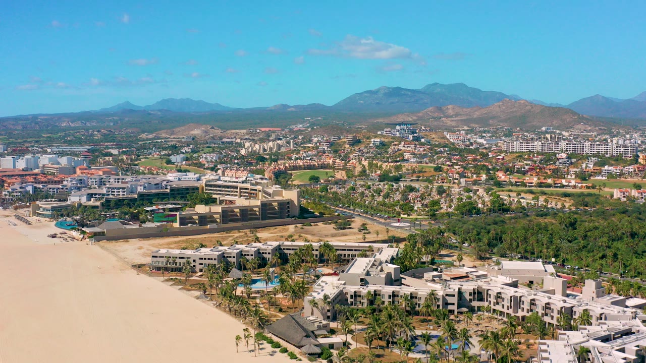 Wide aerial frame over San Jose Del Cabo shows sandy beach at left, low beachfront buildings and palms below, inland roads and development, mountains in the distance, and no visible people.