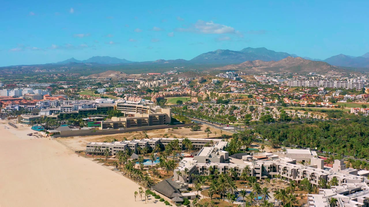 Aerial view in San Jose Del Cabo looks past pale sand and beachfront buildings toward a road grid, low-rise development, dry hills, distant mountains, clear sky, and no visible people.