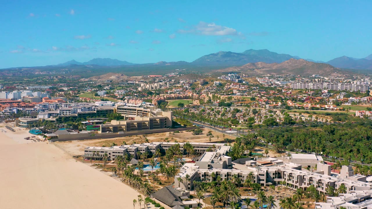 The San Jose Del Cabo aerial frame shows the sandy beach edge, white beachfront structures, palm clusters, town blocks, tan hills, blue mountain horizon, and no visible people.