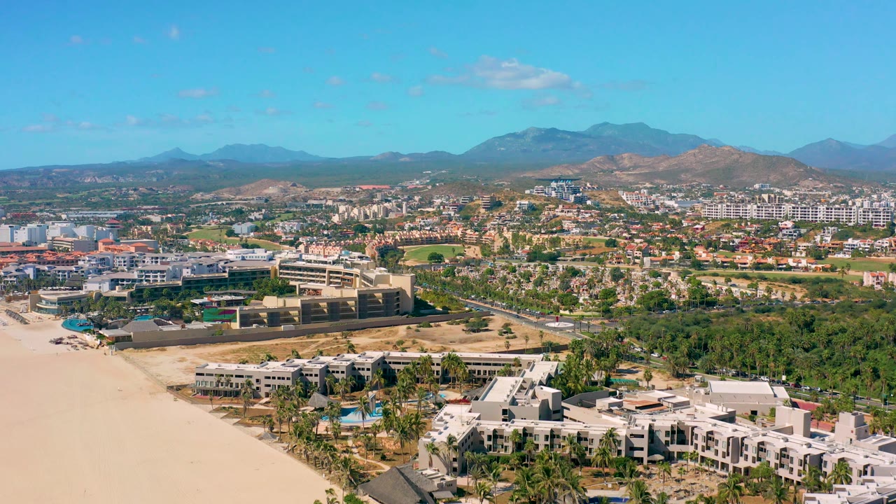 Aerial coastline view with beach, road, buildings, palms, hills, and mountains.