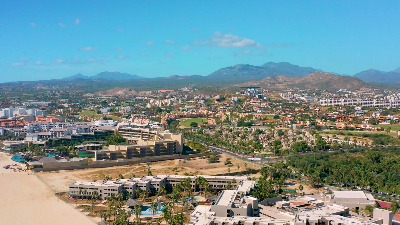 Wide aerial frame of San Jose Del Cabo showing pale sand and rooftops in the foreground, a developed urban area beyond, distant mountains, blue sky, and no visible people.