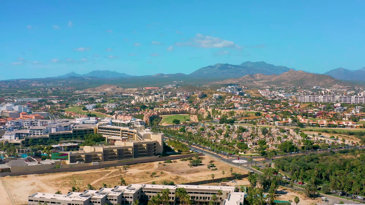 Wide aerial view of urban buildings, palm-lined roads, traffic, dry land, and mountain horizon.