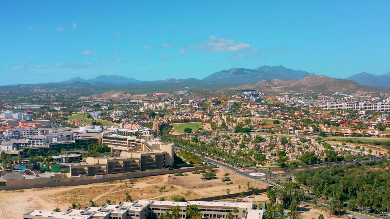 High aerial view in San Jose del Cabo with low-rise development, a road crossing the foreground, dry open lots, green planting, distant hills, and no visible people.
