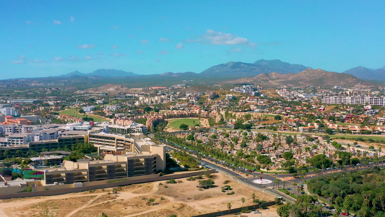 Opening aerial frame over San Jose del Cabo with dry foreground land, a large multi-story building, roads, residential blocks, distant mountains, blue sky, and no visible people.