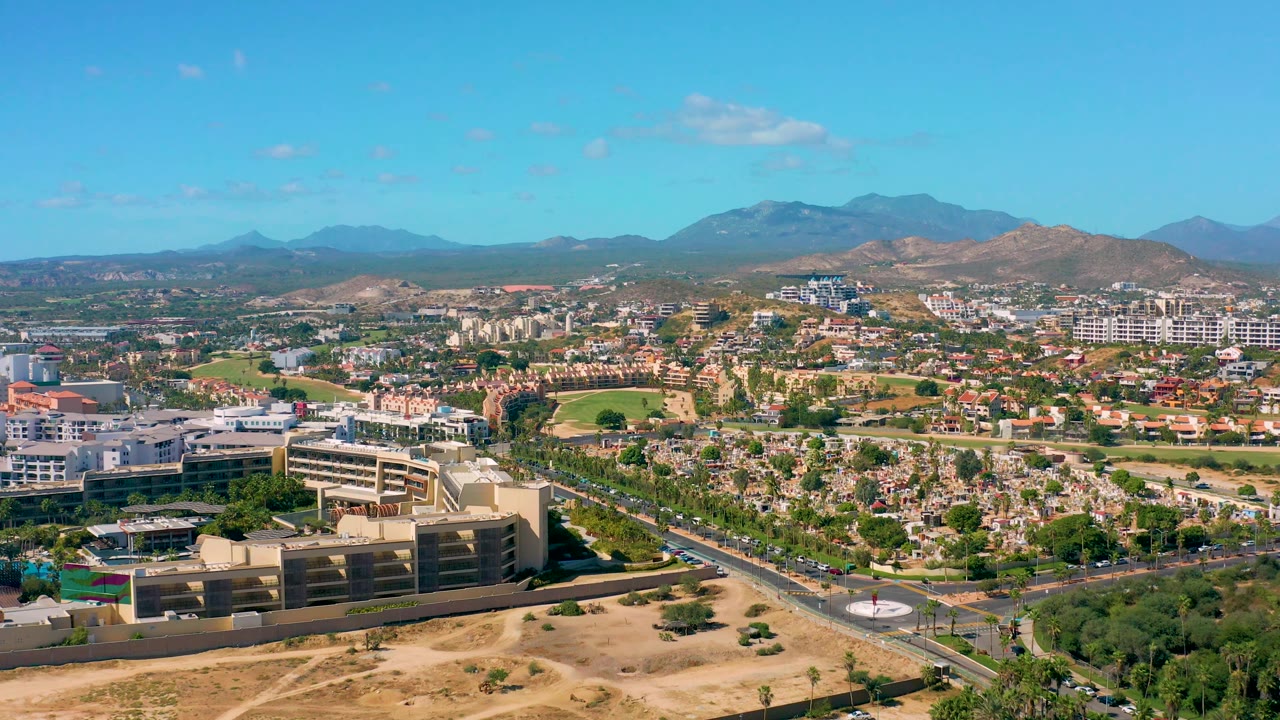 San Jose del Cabo aerial frame with visible traffic on the roads, clustered homes, several larger structures, tan undeveloped ground in front, distant mountains, and no visible people.