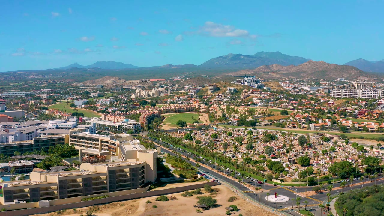 One second into the clip, the San Jose del Cabo aerial view keeps beige foreground buildings, a road with small vehicles, green trees, dry terrain, mountains, blue sky, and no visible people in frame.