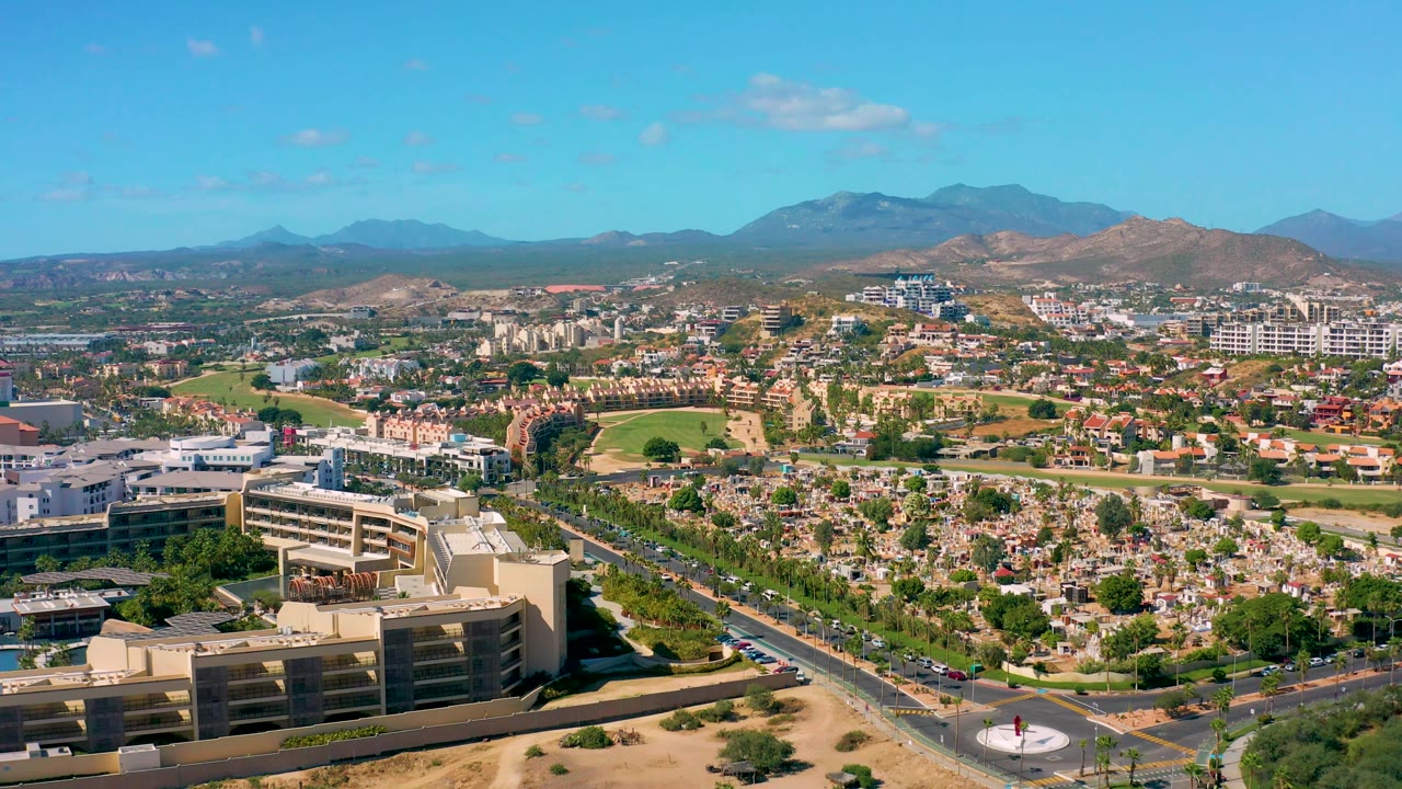 At 0:02, the San Jose del Cabo frame shows the road corridor and foreground development beneath a broad residential area, with dry hills, distant mountains, clear sky, and no visible people.