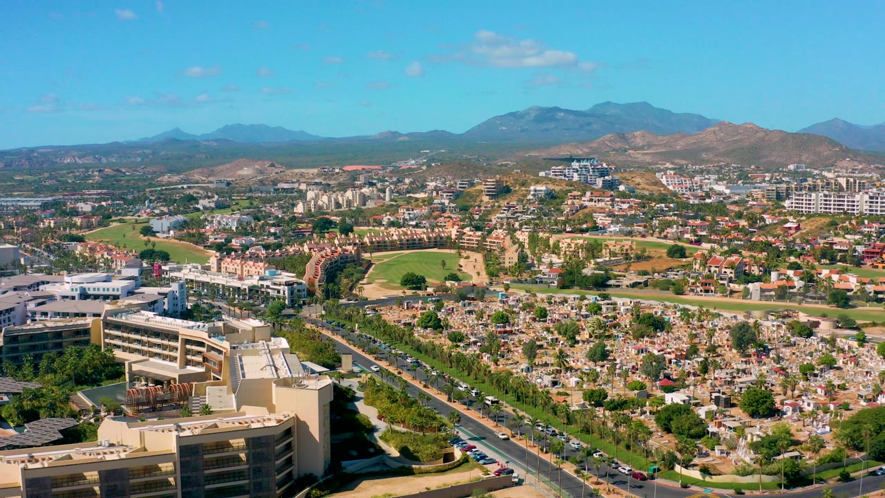 Opening aerial view of San Jose Del Cabo with a large foreground building, a road with cars, urban neighborhoods, green areas, distant mountains, blue sky, and no visible people.