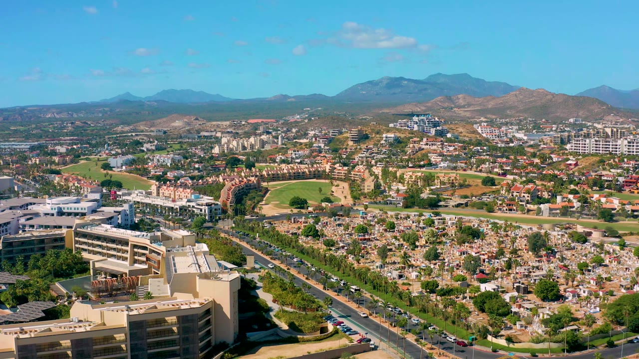 Aerial frame in San Jose Del Cabo showing the road corridor slightly farther through the scene, with cars, low-rise buildings, landscaped green areas, mountains, clear sky, and no visible people.