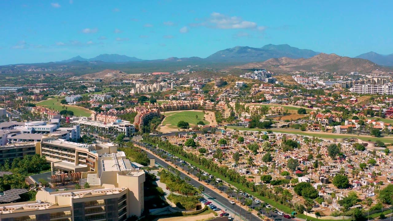 San Jose Del Cabo aerial view with the foreground building lower in frame, a busy road line, clustered homes and commercial buildings, green patches, brown hills, blue mountains, and no visible people.