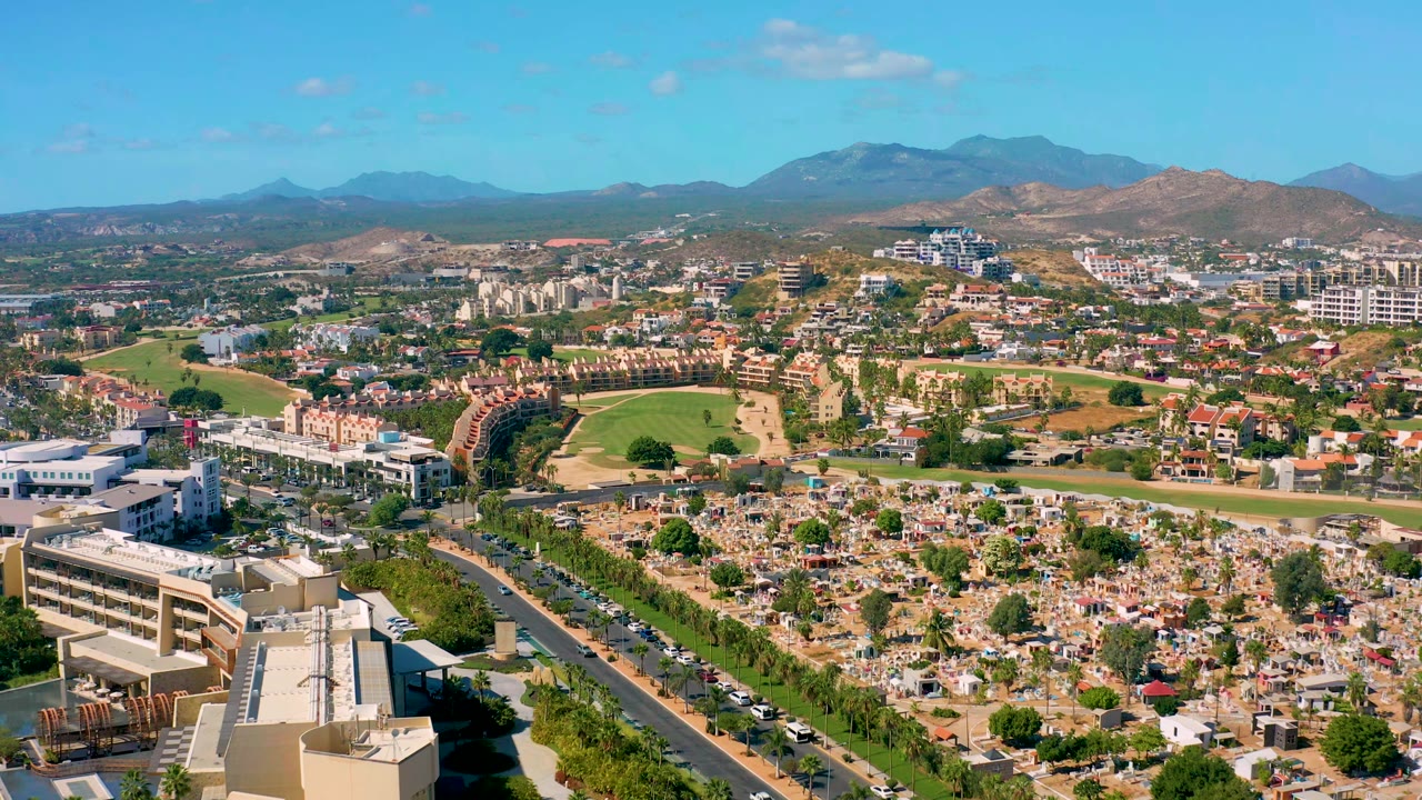 Second frame shows San Jose Del Cabo from above with dense cemetery plots lower in frame, cars on bordering roads, mixed buildings, green fields, and no visible people.