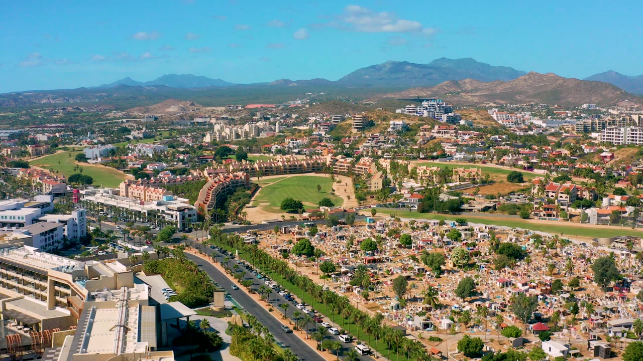 Aerial city view with cemetery foreground, parked and moving cars, green field, and hills.