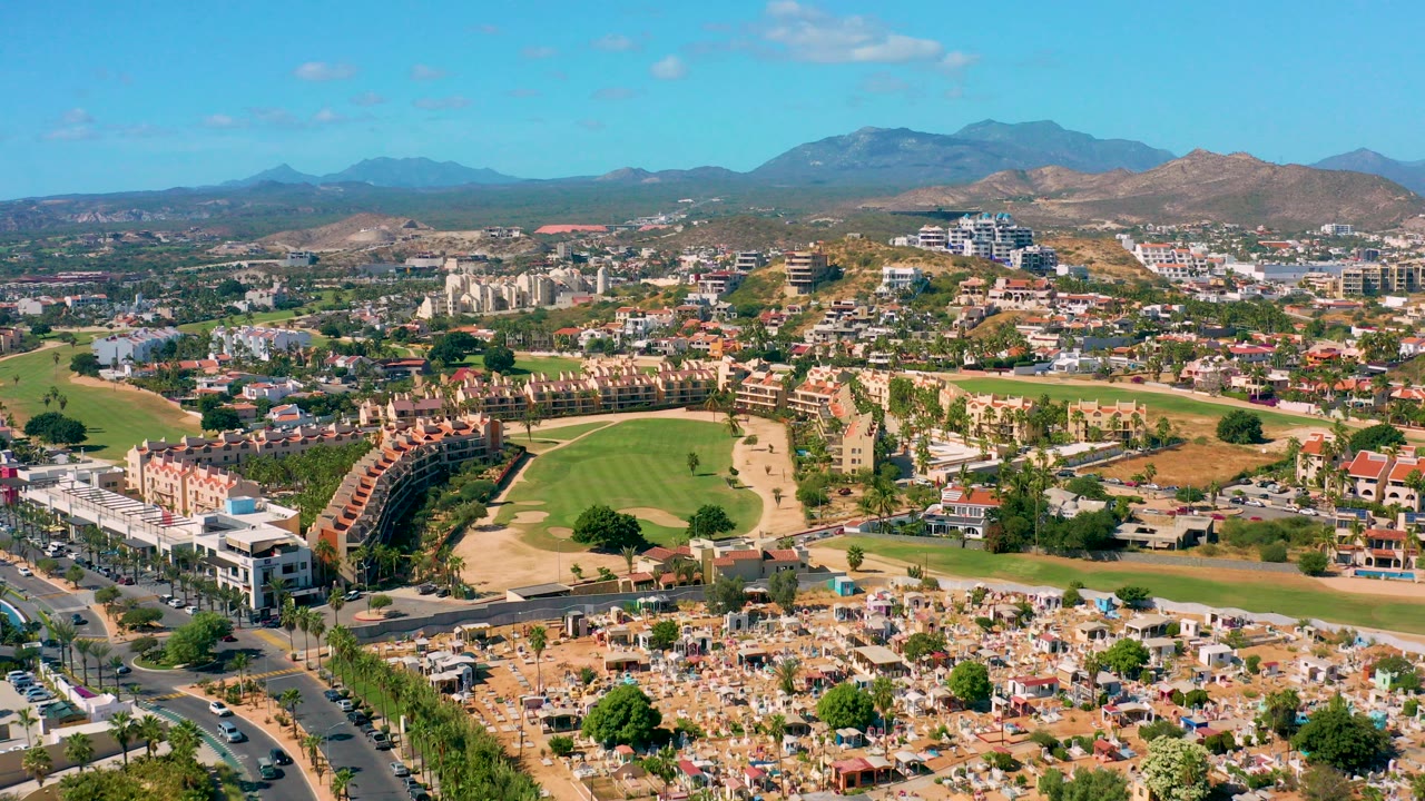 Opening aerial frame over San Jose Del Cabo with a green golf course, curved residential buildings, roads, dry hills, distant mountains, and no visible people.