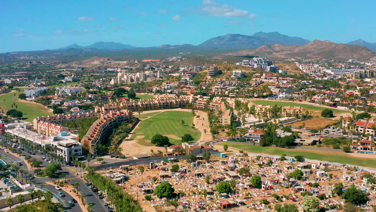 Early pan frame in San Jose Del Cabo showing the golf fairway centered between curved housing, roadside buildings, distant mountains, and no visible people.