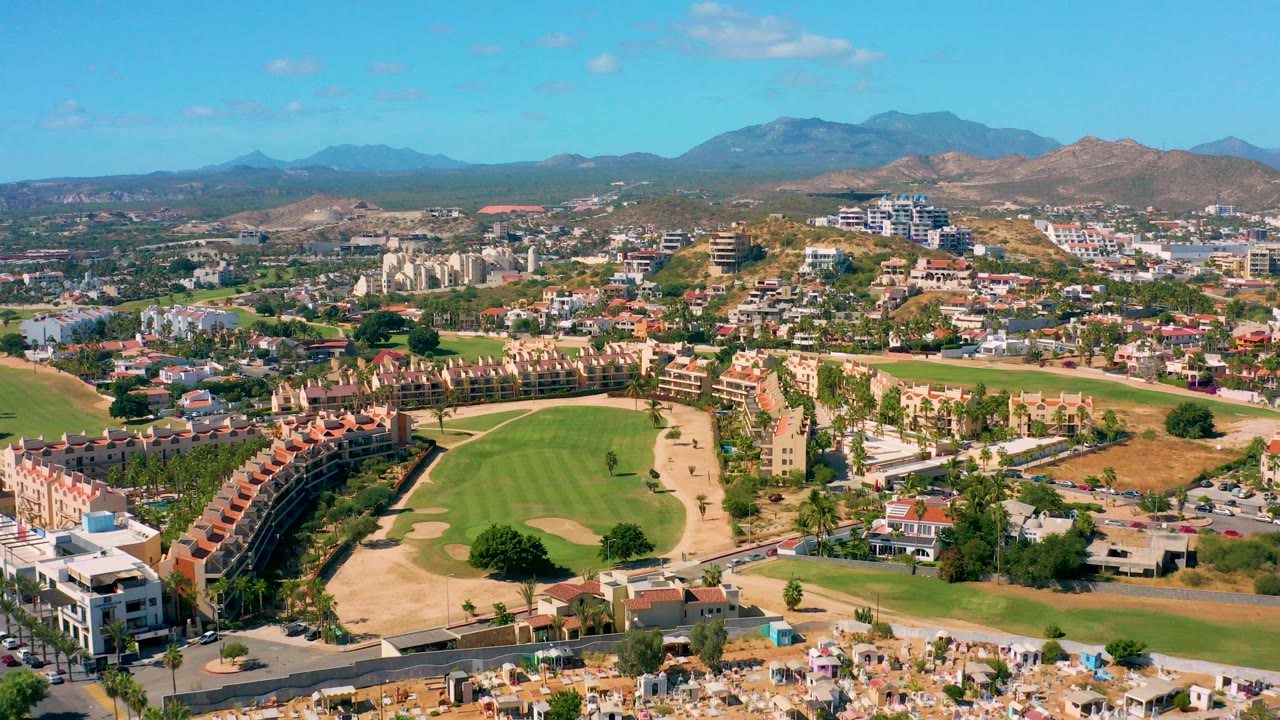 Wide aerial view over San Jose del Cabo with a green golf fairway, sand bunkers, rows of homes, low-rise buildings, distant mountains, blue sky, and no visible people.