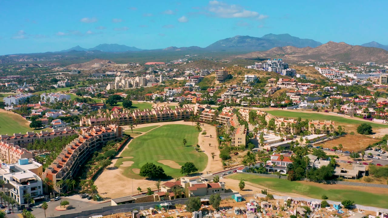 San Jose del Cabo appears from a high drone view with the golf course running across the lower frame, clustered homes and apartment blocks beyond it, dry terrain, mountain ridges, and no visible people.