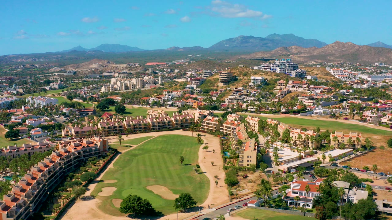 Wide aerial view over a San Jose del Cabo golf course with green fairways, sand traps, red-tiled buildings, distant mountains, blue sky, and no visible people.