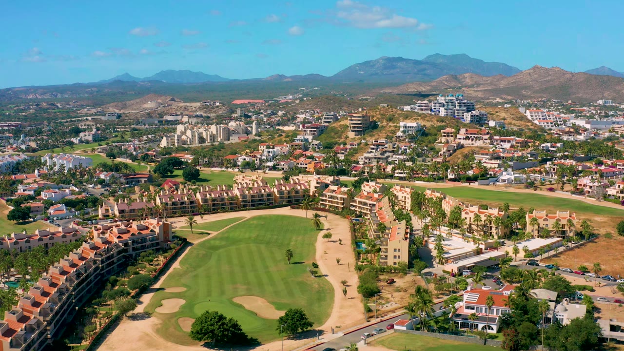 High drone view in San Jose del Cabo showing golf fairways in the foreground, red-roof resort buildings around the course, dry hills, mountains, clear sky, and no visible people.