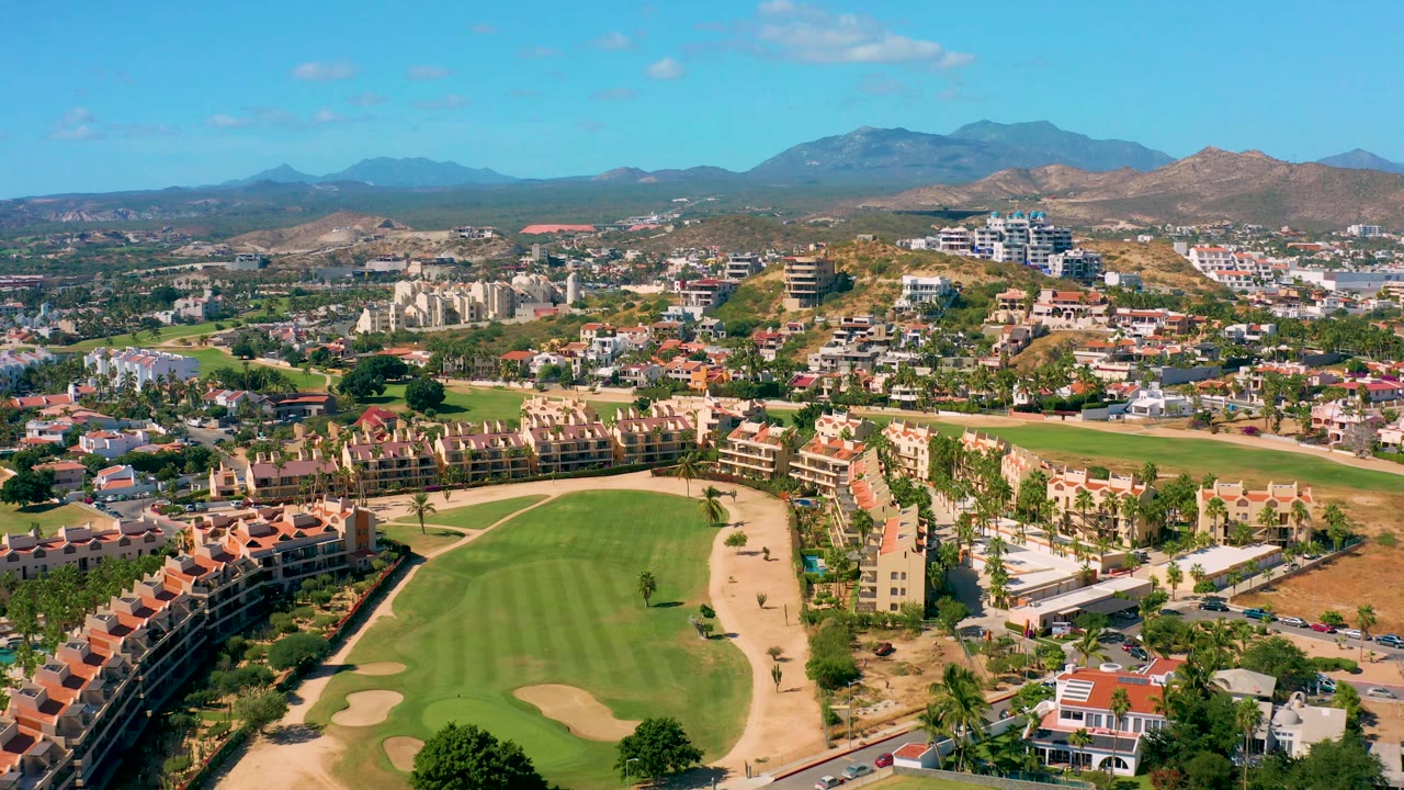 Aerial frame over San Jose del Cabo with the golf course lower in frame, palm-lined development and resort buildings across the middle, mountains beyond, and no visible people.