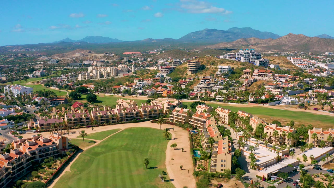Wide aerial view over San Jose Del Cabo showing a green golf course foreground, red roof residential buildings, hillside development, distant mountains, blue sky, and no visible people.